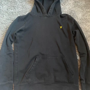 Svart hoodie från Lyle & Scott - Svart hoodie från Lyle & Scott med klassisk gul logga på bröstet. Tröjan har huva, känguruficka och långa ärmar. Perfekt för dig som gillar enkel och stilren streetwear. Mjuk och skön bomullskänsla.
