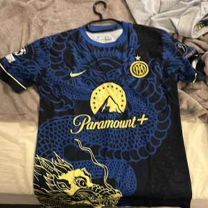 Inter Nike Dri-Fit Calhanoglu tröja M - Inter fotbollströja från Nike med Dri-Fit-material, blå och svart med drakmotiv i gult. Tryck med Calhanoglu 20 på ryggen, samt sponsorloggor från Paramount+ och Lenovo. Kortärmad modell med coolt och unikt mönster, perfekt för fans av Inter.  Pris kan diskutera