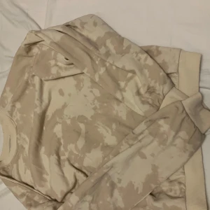 Beige camo sweatshirt från H&M - Säljer en beige sweatshirt från H&M med camouflagemönster i olika ljusa toner. Tröjan har rund hals, ribbade muddar och är långärmad. Perfekt för dig som gillar en chill och avslappnad stil.