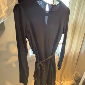 Svart långärmad klänning med knyt - En svart, enkel och stilren klänning med långa ärmar och knytband i midjan. Klänningen har en liten v-ringning framtill och är i kort modell. Perfekt för dig som gillar minimalistisk och klassisk stil. Hör av er om bild på hur den sitter på🫶🏻