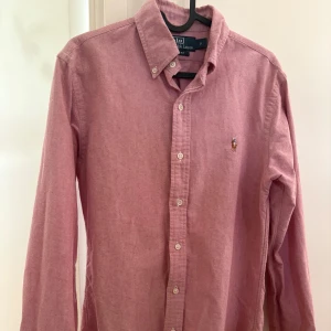 Rosa skjorta från Ralph Lauren - Klassisk rosa skjorta från Ralph Lauren med button-down krage och broderad logga på bröstet. Skjortan har långa ärmar och knäppning framtill. Perfekt för en stilren och avslappnad look.