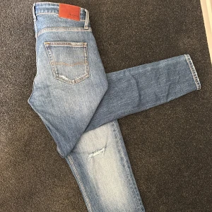 Blå jeans från Pepe Jeans - Säljer ett par blå jeans från Pepe Jeans med slitna detaljer vid knäna. Klassisk femficksmodell med läderpatch bak i midjan och orangea sömmar. Perfekta för dig som gillar en smal passform och lite tuffare look. Aldrig använda!!
