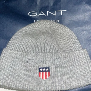 Grå ribbstickad mössa från Gant - Snygg grå ribbstickad mössa från Gant. Klassisk modell med uppvikt kant och diskret design som passar till det mesta. Perfekt för kyliga dagar när du vill hålla stilen enkel men ändå trendig.