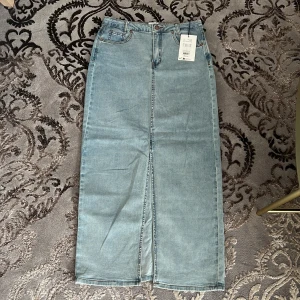 Ljusblå jeanskjol från My Essential Wardrobe - Lång, ljusblå jeanskjol med klassisk femficksdesign och hög midja. Kjolen har en snygg slits framtill som ger extra rörelsefrihet. Perfekt för dig som gillar en clean och tidlös denimlook. Helt oanvända 