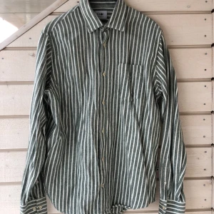 NN07 grönvit skjorta, storlek large - Tjockare grön och vit randig skjorta med långa ärmar och en bröstficka. Skjortan har en klassisk krage och knappar framtill samt vid manschetterna. Från No Nationality. Använd 2-3 gånger, helt i nyskick.