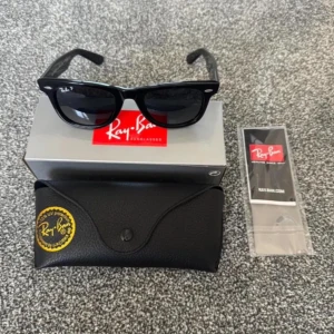 Svarta Wayfarer solglasögon från Ray-Ban - Klassiska svarta Wayfarer solglasögon från Ray-Ban med glansig finish och tydlig logotyp på bågen. Modellen har en tidlös, fyrkantig form och levereras med originalfodral och putsduk.