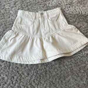 Vit volangkjol i jeansmaterial - Supersöt vit kjol i jeansmaterial med volang nedtill och klassiska fickor fram. Kjolen har knapp och dragkedja i midjan och en lätt utsvängd form. Perfekt att styla med en enkel topp eller hoodie.