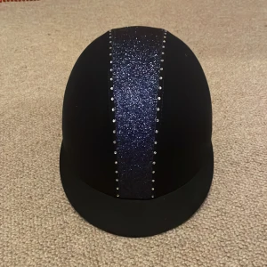 mörkblå  glittrig ridhjälm med stenar - Snygg svart ridhjälm med bred glittrig mörkblå rand framtill och små glittrande stenar längs sidorna. Hjälmen har klassisk form och medföljer en svart dragskopåse för förvaring. Perfekt för dig som vill sticka ut i stallet. Hjälmen är använd fåtal gånger och är i nytt skick!