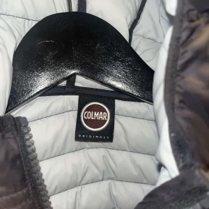 Svart Colmar pufferjacka med huva - Säljer en svart pufferjacka från Colmar Originals med huva och dragkedja. Jackan har quiltad design, två fickor med dragkedja och Colmar-logga på ärmen. Perfekt för dig som vill ha en stilren och varm jacka med snygg passform. Storlek 10y 142 cm