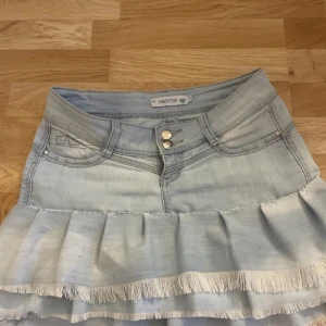 Ljusblå volangkjol från Mantis - Supersöt ljusblå jeanskjol från Mantis med volangkant och fransiga detaljer. Kjolen har klassiska jeansfickor fram och bak, dubbla knappar och en snygg metallbricka bak. Perfekt för dig som gillar en cool och feminin stil.