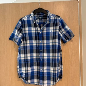 Blårutig kortärmad skjorta Ralph Lauren - Kortärmad skjorta från Ralph Lauren i blått, vitt och grått rutigt mönster. Klassisk button-down krage, broderad logga på bröstet och knäppning framtill. Tillverkad i mjuk bomull, perfekt för en chill och stilren look.
