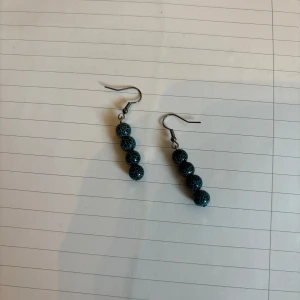 Svarta och blå glittriga örhängen med pärlor - Säljer ett par snygga örhängen med tre runda pärlor i svart och blått med glittrig yta. Hängande modell med metallkrok. Perfekta för att ge din outfit lite extra färg och glans!