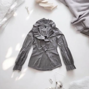 Grå blus med volangkrage - Säljer en grå blus med stor volangkrage och långa ärmar med rynkade muddar från Stile Benneton. Blusen har en figurnära passform och subtilt rutigt mönster. Perfekt för dig som vill sticka ut med en trendig och elegant look.