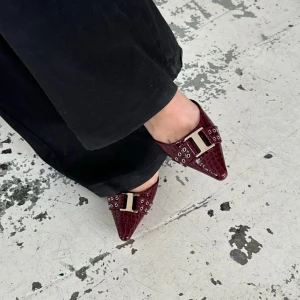 Vinröda pumps med krokodilmönster och spänne - Säljer ett par vinröda pumps med spetsig tå och krokodilmönstrad yta. Skorna har en dekorativt stort metallspänne framtill och justerbar rem runt hälen. Perfekta för dig som vill sticka ut med en elegant och trendig look. Säljs pågrund av att dem är för stora för mig och är alldeles för dyrt att skicka tillbaka till hemsidan. Aldrig använda med det sagt.