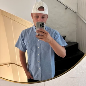 Blå kortärmad skjorta från Lacoste - Snygg ljusblå skjorta från Lacoste med korta ärmar och klassisk button-down-krage. Skjortan har en bröstficka med den ikoniska krokodil-loggan och vita knappar framtill. Perfekt för en clean och avslappnad stil.