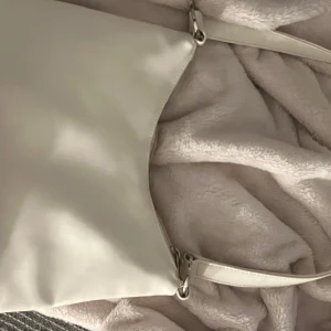 Vit väska  - Säljer en stilren vit axelväska med justerbart axelband och silvriga metalldetaljer. Väskan har dragkedja upptill och flera innerfack för att hålla ordning på dina grejer. Perfekt för dig som gillar minimalistisk och clean look.