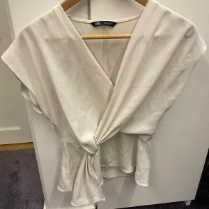 Vit omlottopp från Zara - Snygg vit omlottopp från Zara med knyt fram och v-ringad hals. Toppen har korta ärmar och en lätt glansig yta som ger en stilren look. Perfekt att matcha med jeans eller kjol för en trendig outfit.