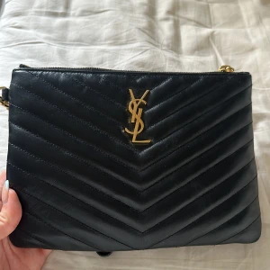 Svart YSL kuvertväska i skinn - Säljer en svart kuvertväska från YSL i quiltat skinn med guldiga detaljer och dragkedja upptill. Väskan har det klassiska YSL-emblemet framtill och ett stilrent, elegant mönster. Den är som ny och har inga defekter på sig. Den är köpt för 8500kr på ysl hemsida. Har kvitto och box kvar 