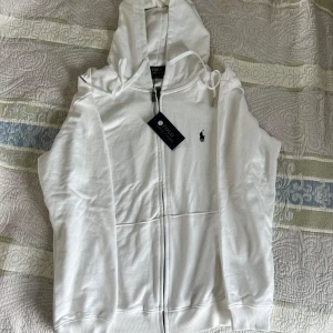 (Helt ny) Vit hoodie från Polo Ralph Lauren - Snygg vit hoodie från Polo Ralph Lauren med dragkedja och huva. Klassisk broderad logga på bröstet och ribbade muddar. Den är endast testats men är för stor för mig… alltså helt ny. Påse och etiket får med. Obs (k@pia)