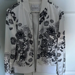 Vit bomberjacka med drakmönster från Guess - Snygg vit bomberjacka från Guess med svart drak- och blommönster över hela jackan. Jackan har dragkedja framtill, ribbade muddar och är köpt i guess miami . Perfekt statement-plagg för dig som vill sticka ut. Jackan är XS men passar S med