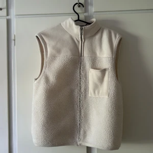 Beige fleeceväst med dragkedja - Mysig beige fleeceväst med hög krage. Västen har en enkel bröstficka och är tillverkad i mjukt teddyfleece-material. Perfekt lager-på-lager-plagg för kyliga dagar. Använd 1 gång och i väldigt fint skick. Nypris 500