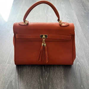 Orange handväska med tofsdetalj - Snygg orange handväska i skinnimitation med guldfärgade metalldetaljer och dekorativa tofsar framtill. Väskan har ett stadigt handtag och medföljande axelrem för extra flexibilitet. Perfekt storlek för att få plats med allt du behöver och flera fack inuti.