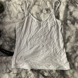 Vit blommig linnetopp från Winner - Supersöt vit linnetopp med tunna axelband dekorerade med virkade blommor och pärldetaljer. Toppen har v-ringning och små blommor broderade framtill för en extra gullig look. Perfekt för varma dagar och somriga outfits! Det står det är för M fast det passar mig perfekt som bär Xs/S 