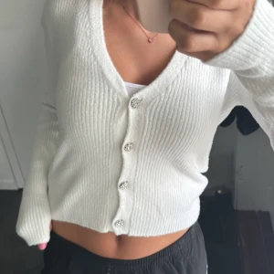 Vit ribbad kofta från Zara - Superfin vit ribbad kofta från Zara med glittriga blomformade knappar framtill. Croppad modell med v-ringning och långa ärmar, perfekt att styla med jeans eller kjol. Mjuk stickad känsla som gör den extra skön att ha på sig.