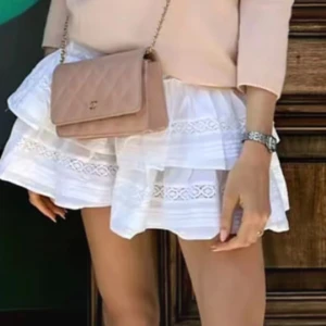 Vit volangkjol med spetsdetaljer - Supersöt vit kortkjol med bred smock i midjan och flera lager volanger. Kjolen har fina spetsdetaljer och broderier som ger en romantisk vibe. Perfekt för sommarens alla tillfällen och lätt att matcha med en enkel topp.