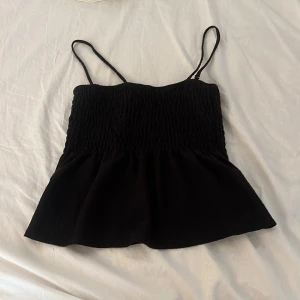 Svart smockad linnetopp - Supersöt svart linnetopp med smockad överdel och tunna axelband. Toppen har en lätt utsvängd nederdel som ger en flowy känsla. Perfekt att styla med jeans eller kjol. 