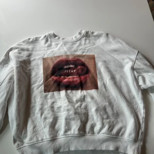 Vit sweatshirt från ISTAY med tryck - Säljer en vit sweatshirt från ISTAY i storlek L. Tröjan har ett stort tryck med röda läppar och texten 'ISTAY' på framsidan. Rund hals och ribbade muddar vid ärmslut och nederkant. Perfekt för dig som gillar statement-plagg och streetstyle.