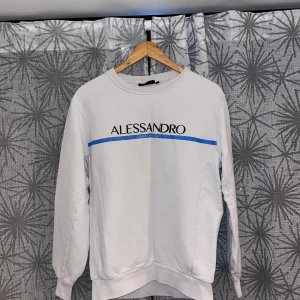 Vit Alessandro sweatshirt med blå detalj - Snygg vit sweatshirt från Alessandro med lång ärm och rund hals. Framsidan har ett svart tryck med märkesnamnet och en blå linje som går tvärs över bröstet. Tröjan är gjord i mjuk bomull och har ribbade muddar vid ärmslut och nederkant. Perfekt för chill dagar.