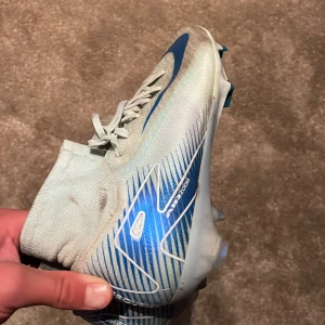 Nike Air Zoom fotbollsskor blå/vit - Säljer ett par Nike Air Zoom fotbollsskor i vitt med blå detaljer och snörning. Skorna har en strumpliknande passform och mönstrad ovandel i syntetmaterial. Sulan har blå och vita inslag med dobbar för gräsplan. Perfekt för dig som vill ha grymt grepp och snygg stil på planen.
