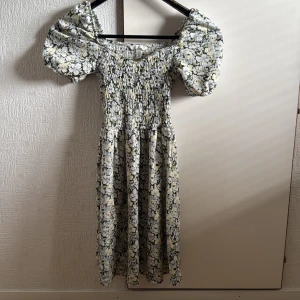Blommig offshoulder klänning XS - Supersöt blommig klänning från H&M i storlek XS. Offshoulder med puffiga korta ärmar och smock upptill. Mönstret är blommigt i vitt, svart och ljusgult. Kjolen är lätt utställd och klänningen är knälång. Perfekt för sommarens alla tillfällen! 