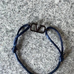 Valentino blått armband med V-logo - Snyggt armband från Valentino med justerbart blått snöre och en metallfärgad V-logo i mitten. Perfekt accessoar för att lyfta din stil med en touch av lyx. Diskret metallbricka med Valentino-gravyr vid knuten.