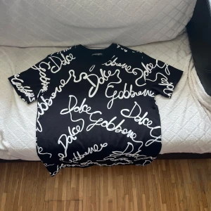 Svart Dolce & Gabbana t-shirt med print - Svart t-shirt från Dolce & Gabbana med vit, handskriven logoprint över hela plagget. Rund hals och korta ärmar. Tillverkad i mjuk bomull som känns skön mot huden. Perfekt för dig som gillar statement-plagg och vill sticka ut med en lyxig vibe. Begränsad utbud, kan ej hittas av denna design längre. Har dock ej originalförpackning eller tagg.