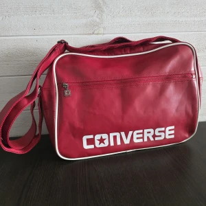 Röd axelväska från Converse - Snygg röd axelväska från Converse med vit piping och stort logotryck på framsidan. Väskan har justerbar axelrem, flera fack med dragkedja och coolt stjärndetalj på zip-pullern. Perfekt för att bära med sig allt man behöver till skolan eller stan.
