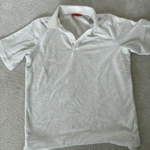 Vit pikétröja från RYVLS - Vit pikétröja från RYVLS i klassisk design med krage och två knappar vid halsen. Tröjan har korta ärmar och är gjord i mjuk bomull som känns skön mot huden. Perfekt för dig som gillar enkel och stilren look.