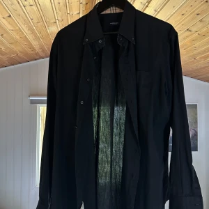 Svart Burberry skjorta i bomull - Svart skjorta från Burberry med klassisk krage och knappar framtill. Långa ärmar med den ikoniska Burberry-rutiga insidan på manschetten. Tillverkad i lätt och luftig bomull, perfekt för dig som gillar stilrena plagg. Köpt på vestaire
