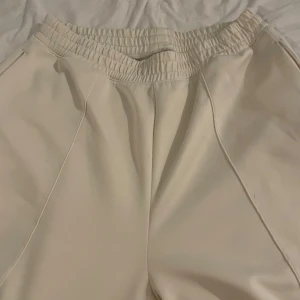 Beiga mjukisbyxor med dragkedja - Säljer ett par beiga mjukisbyxor med elastisk midja och dragkedja vid fickorna. Byxorna har raka ben och är sydda i ett mjukt, syntetiskt material. Perfekta för chill dagar eller när du vill ha det bekvämt.