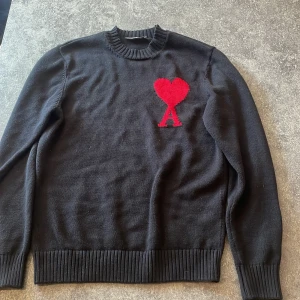 Svart stickad tröja med rött hjärta - Svart stickad tröja från AMI Paris med ett stort rött hjärta och bokstaven A broderat på bröstet. Tröjan har rund hals, ribbade muddar och lång ärm. Perfekt för dig som gillar stilrena plagg med en twist.