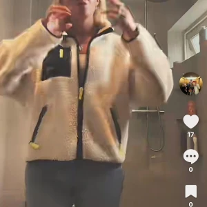 Beige teddyjacka med svarta detaljer - Säljer en beige teddyjacka med svarta detaljer och dragkedjor. Jackan har en oversized passform, hög krage och flera fickor med svarta och gula dragkedjor. Perfekt för dig som vill ha en mysig och trendig look under kyliga dagar.