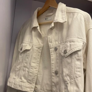 Vit jeansjacka från Gina  - Snygg vit jeansjacka från gina i klassisk modell med krage, bröstfickor med knappar och silvriga detaljer. Jackan har en rak passform och är tillverkad i kraftig bomull. Perfekt att slänga över en hoodie eller t-shirt.