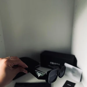 Svarta solglasögon från Prada - Snygga svarta solglasögon från Prada med breda skalmar och markerad rektangulär form. Glasögonen har en stilren look med logga på sidan och mörka linser. Perfekta för dig som vill ha en lyxig och trendig accessoar till sommaren.