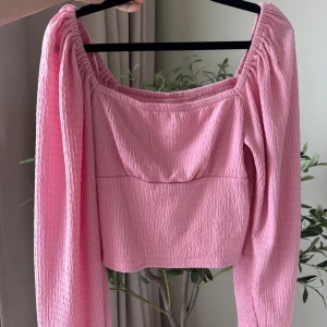 Rosa croppad blus från BikBok - Supersöt rosa croppad blus från BikBok med lång puffärm och smockad struktur. Toppen har en fyrkantig ringning och är gjord i ett lätt, texturerat tyg som ger en cool och trendig vibe. Perfekt att styla med höga jeans eller kjol. OANVÄND 
