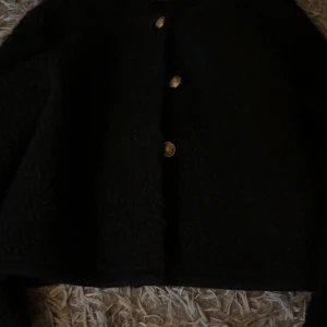 Svart teddyjacka från Gina Tricot - Mysig svart teddyjacka från Gina Tricot i storlek S. Jackan har en fluffig yta, rund hals och snygga guldfärgade knappar framtill. Perfekt för dig som gillar en stilren look med lite extra edge.