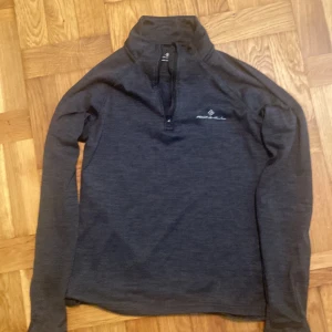 Mörkgrå idrottströja från Ronhill - Mysig mörkgrå långärmad idrottströja med fleece från Ronhill med half zip och hög krage. Perfekt för kyliga dagar eller löprundan. Tröjan har en enkel och sportig look med diskret logga på bröstet och mjukt, borstat foder på insidan. Pris kan diskuteras.