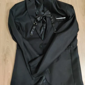 Svart kostym med rosett och kavaj - Stilren svart kostym med klassisk kavaj, matchande byxor och snygg rosett med band. Kavajen har smala slag, knappar framtill och fickdetaljer. Byxorna är raka och enkla. Materialet känns som polyester, perfekt för dig som gillar en clean och elegant look.