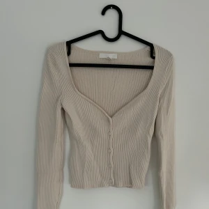 Beige ribbad topp med knappar H&M - Superfin croppad topp från H&M i beige ribbat material. Djup v-ringning, långa ärmar och knappar framtill ger en snygg och trendig look. 