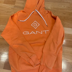 Orange hoodie från GANT med logga - Säljer en snygg orange hoodie från GANT med stor vit logga på bröstet och klassisk huva med vit snörning. Tröjan har ribbade muddar och en skön passform, perfekt för chill dagar. Ficka framtill för mobilen eller nycklar.